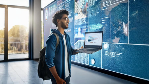 A man with a laptop stands in front of a large digital display showing data.