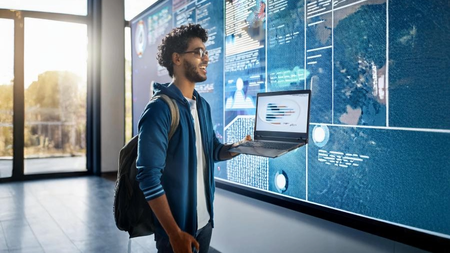 A student with a laptop stands in front of a large digital display showing data.