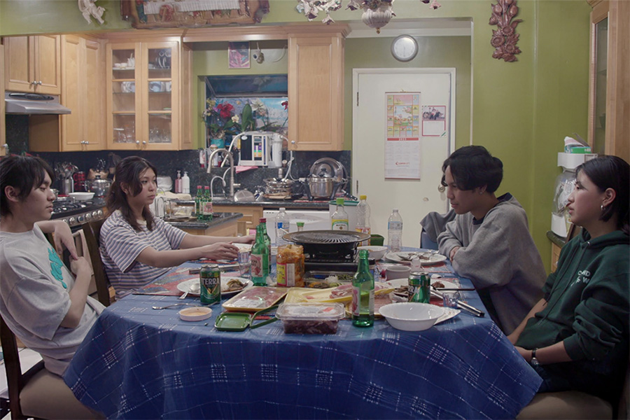 Four young adults sit around a kitchen table looking uncertain. The table is strewn with packages of raw meat and a table-top grill. 