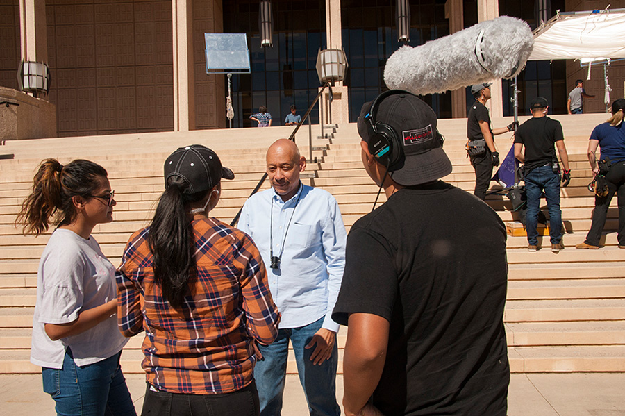 Three young adults, one holding a mic boom, face away from the camera, gathered around a man standing on steps.