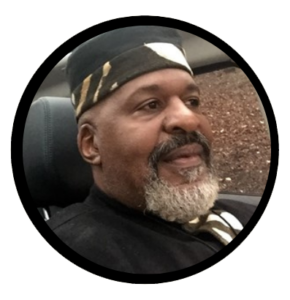 Fluke Fluker wearing a black t-shirt and black bandana on his head. His head is angled and he is looking to his left (your right)