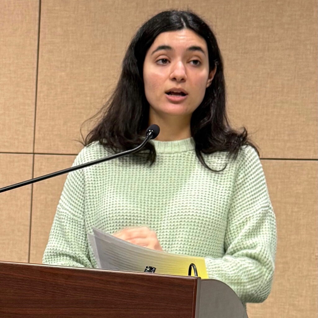 Dark-haired woman in green sweater speaking at a podium