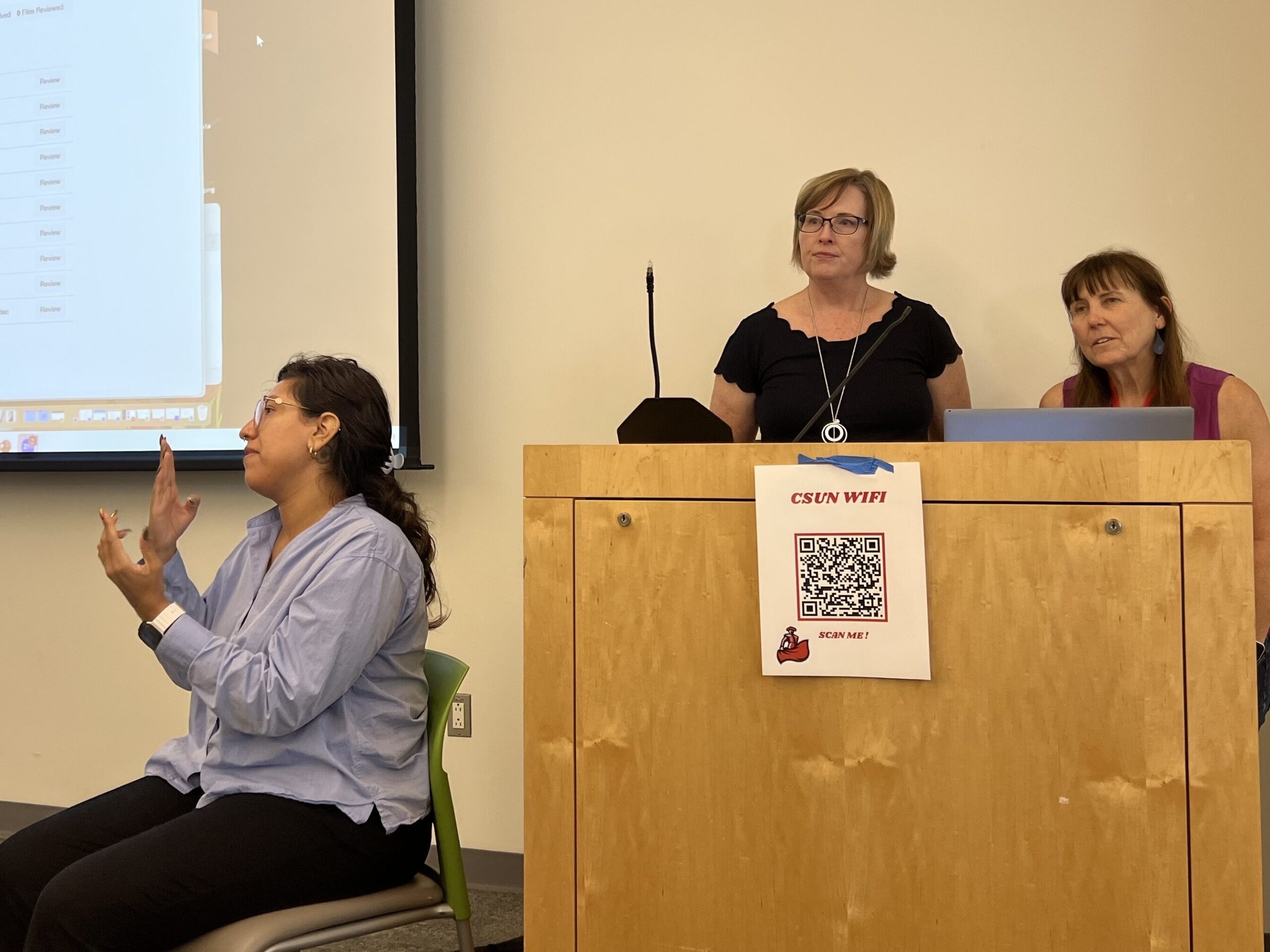 Three people in a classroom setting, one signing and two at a podium, with a projected screen displaying a webpage.