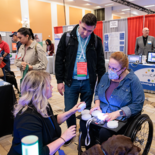 A woman in a wheelchair interacts with a lighted device, assisted by a demonstrator, at a busy technology expo.