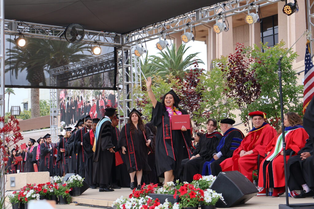 Student walking across stage at commencement ceremony holding one hand up waving to crowd and smiling.