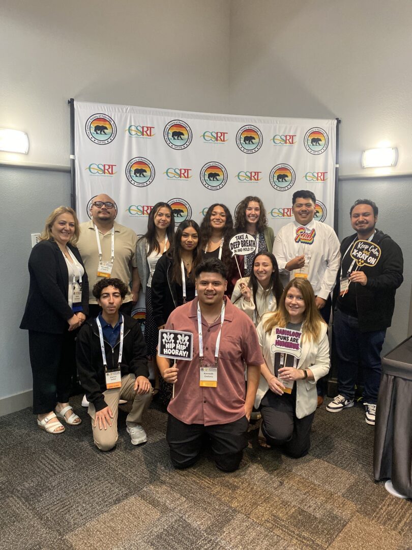A group of CSUN radiologic sciences students and faculty pose at the CSRT Annual Conference in front of a CSRT logo backdrop, holding humorous radiology-themed signs.